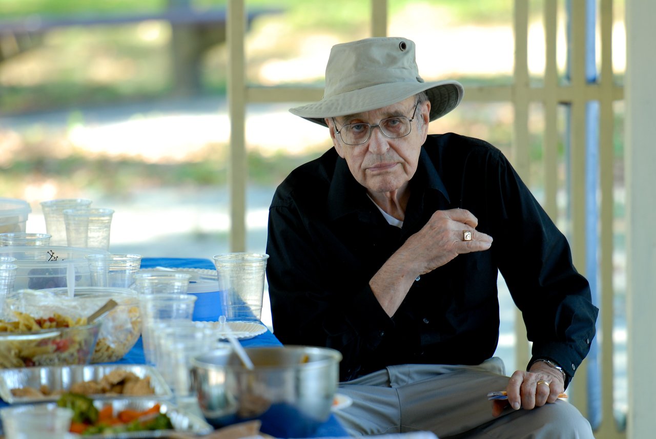 An older man in a hat and glasses sits at a table with food and drinks, looking serious.