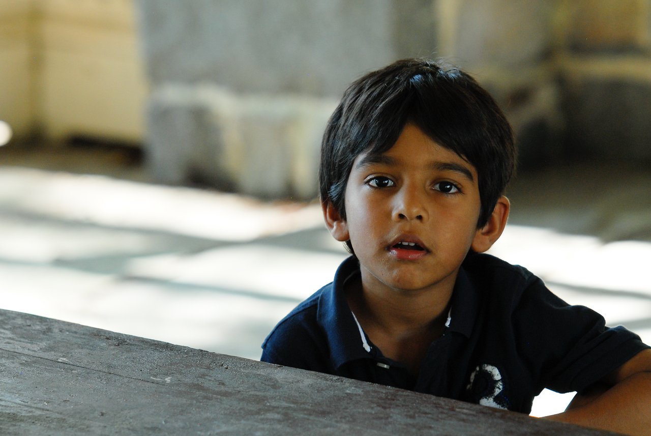 A young boy in a dark shirt sits at a table, looking ahead with a curious expression.
