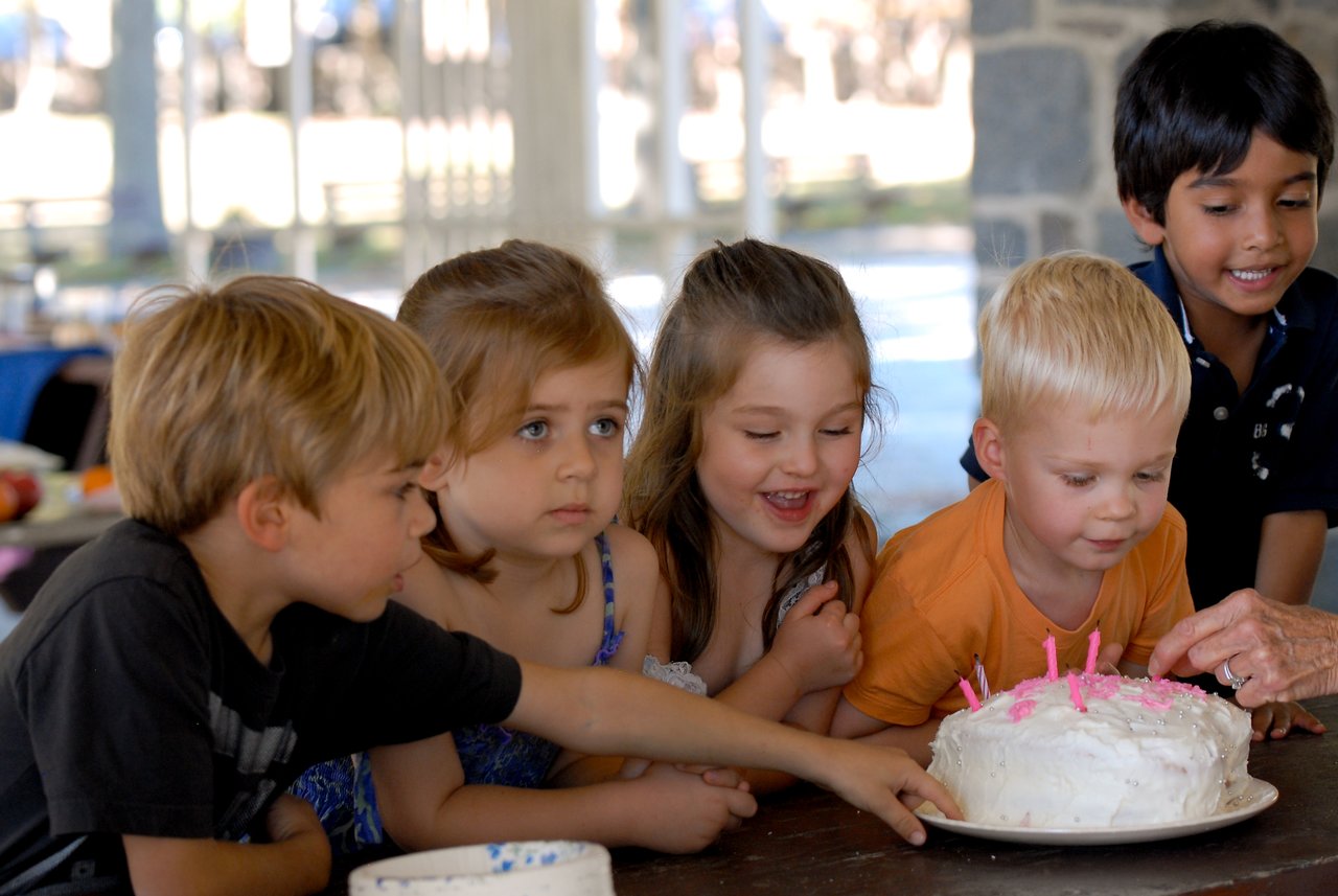 A group of children gathers around a birthday cake with pink candles, excited and reaching toward it.