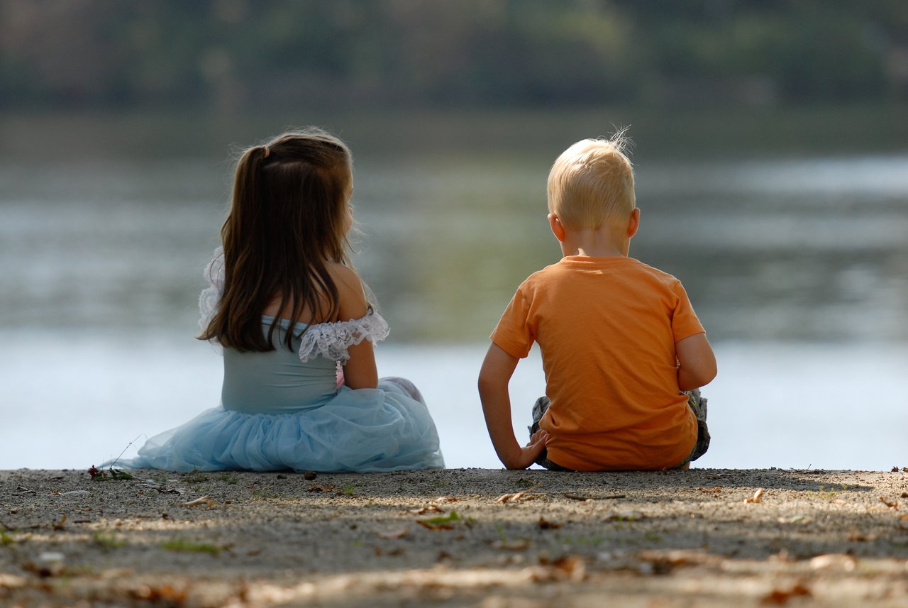 A young girl in a blue dress and a boy in an orange shirt sit together by the water.