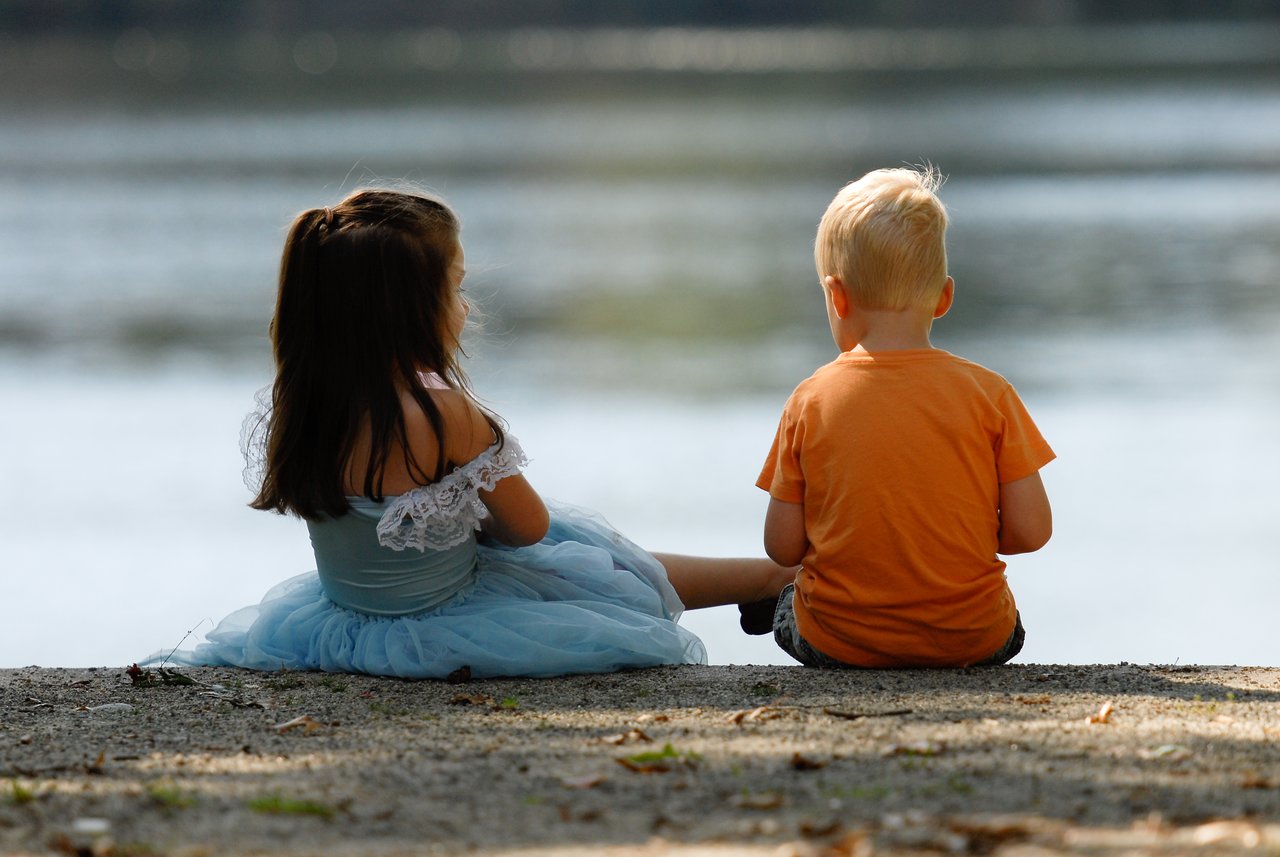 A young girl in a blue dress and a boy in an orange shirt sit by the water together.