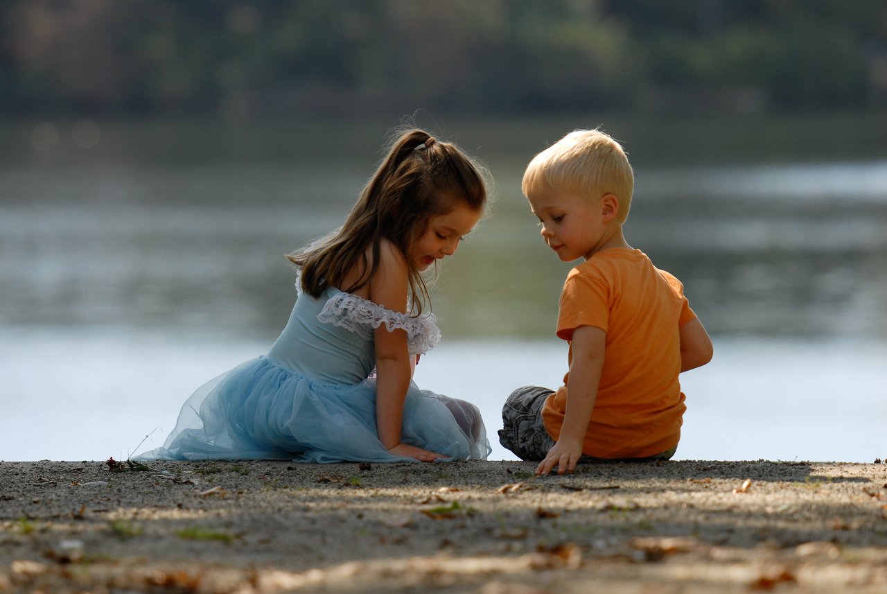A young girl and boy sit on the ground near the water, looking down and interacting quietly.