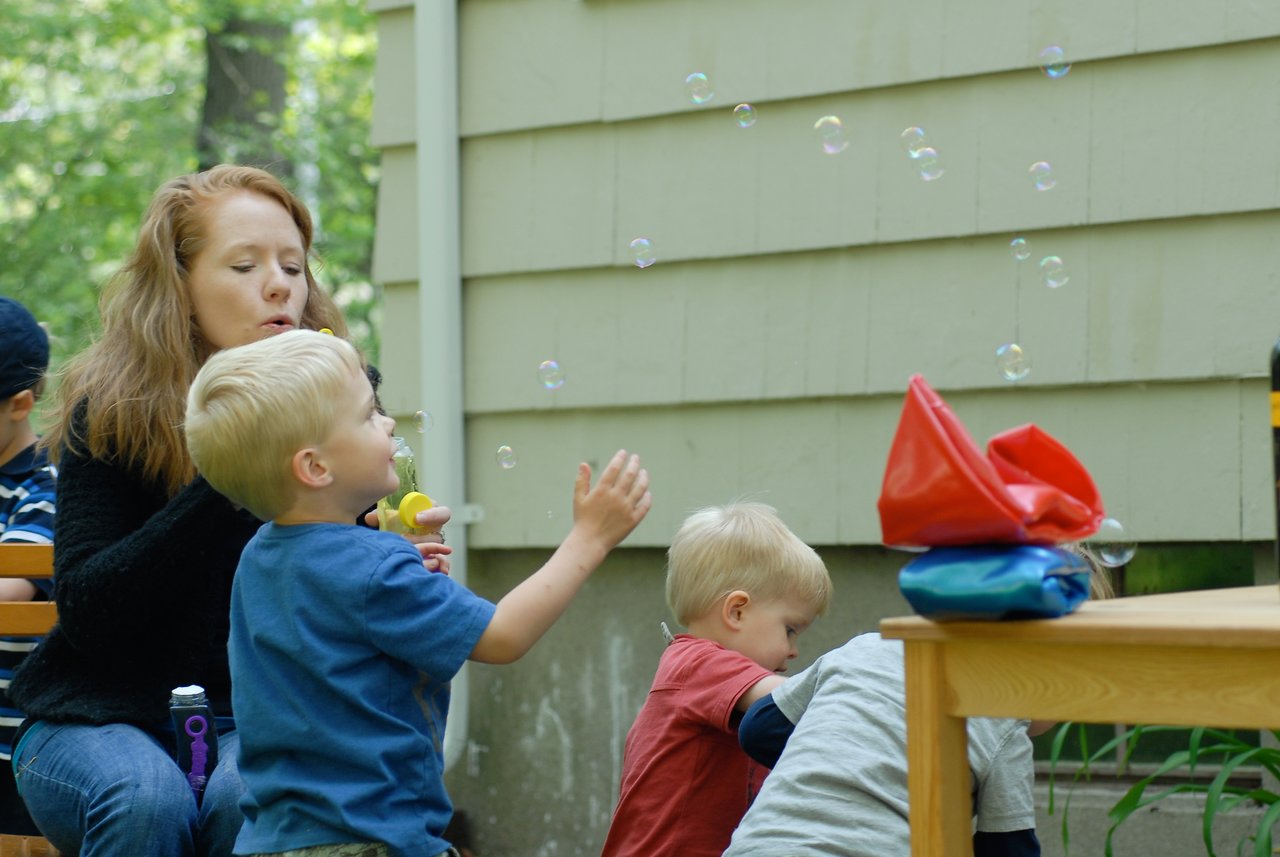 A woman blows bubbles while a young child reaches for them at an outdoor gathering.