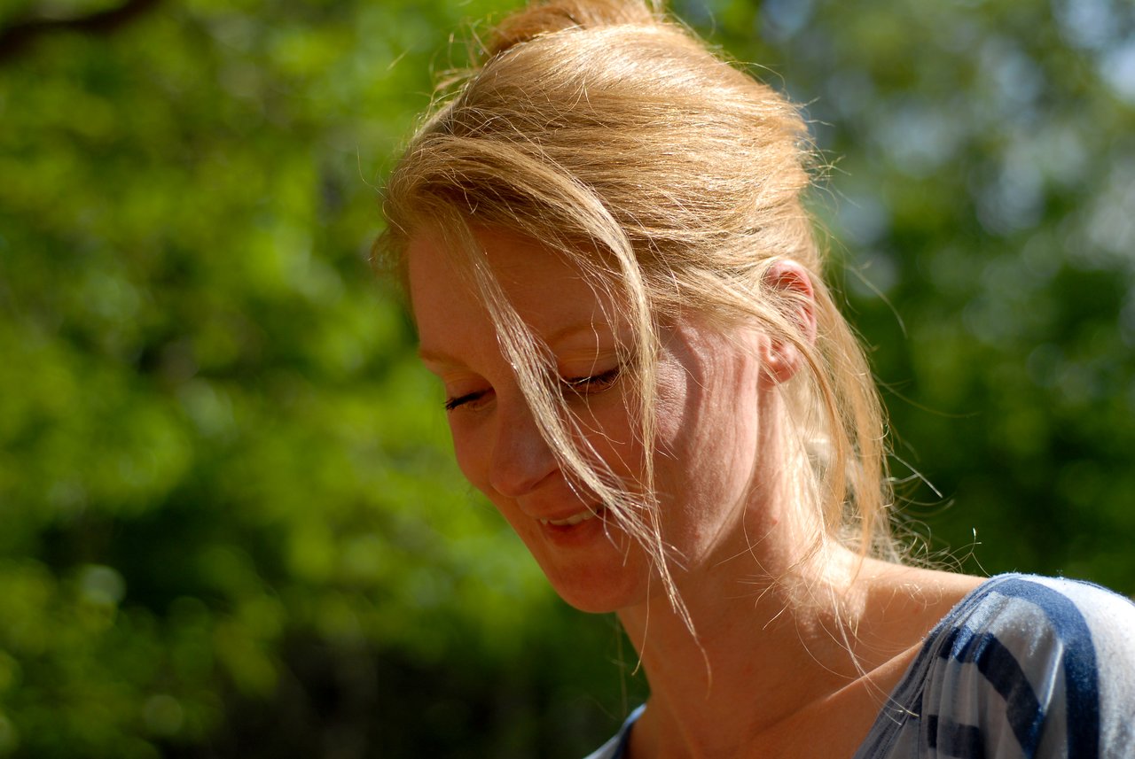 A woman with light skin and blonde hair smiles while looking down at something during an outdoor gathering.