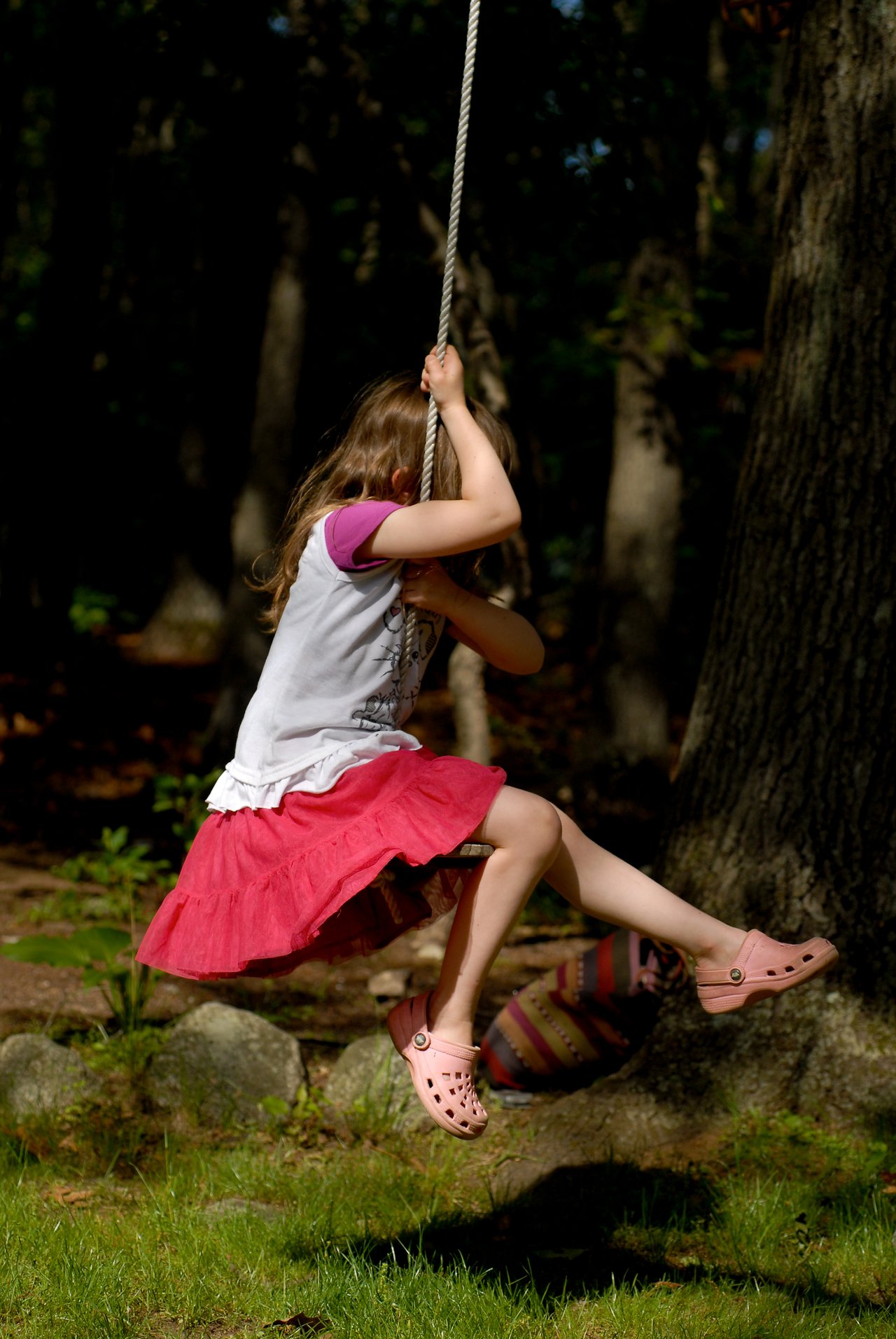 A child in a pink skirt swings on a rope outdoors, holding on tightly with both hands.