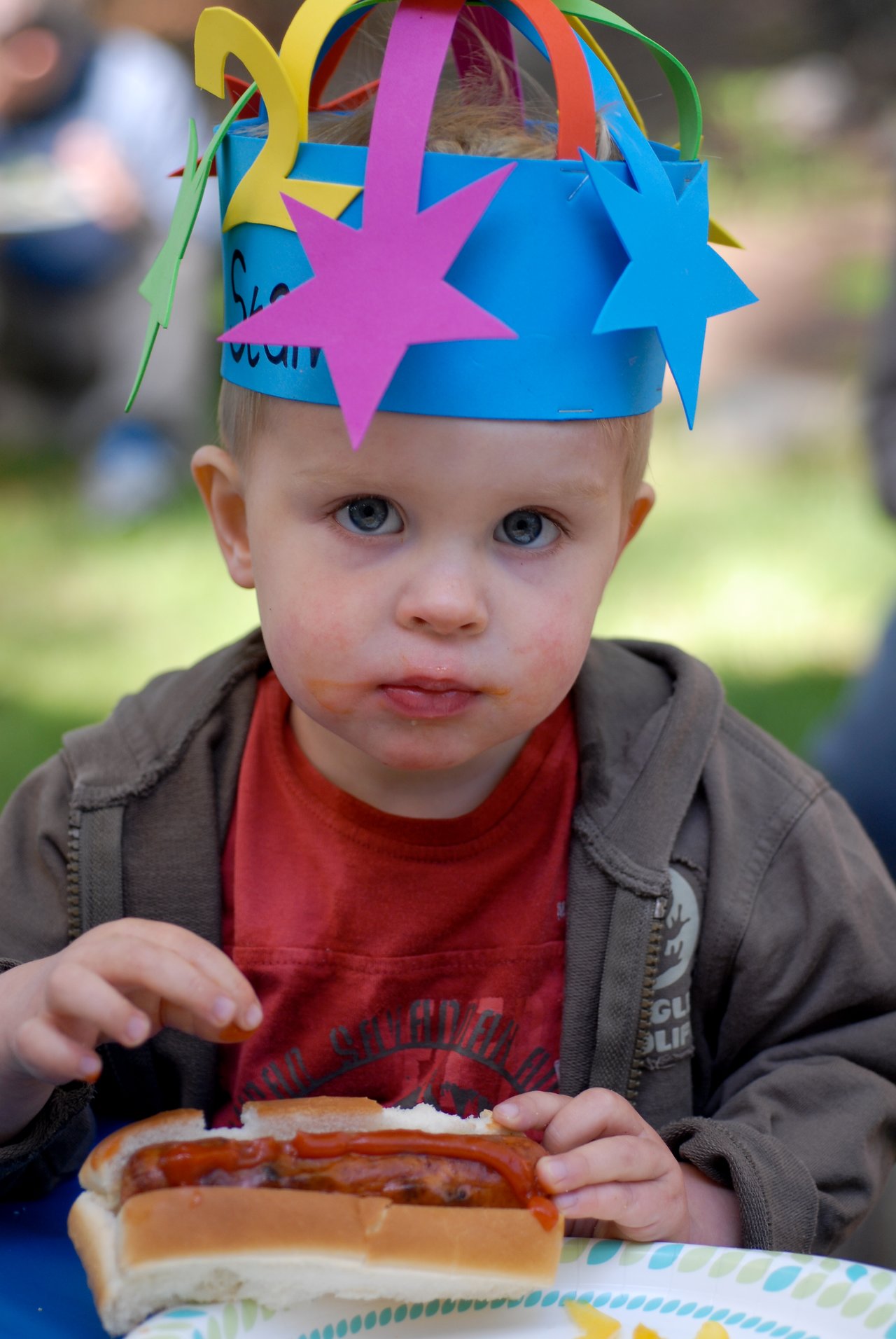 A young child wearing a colorful birthday hat eats a hot dog at an outdoor party.