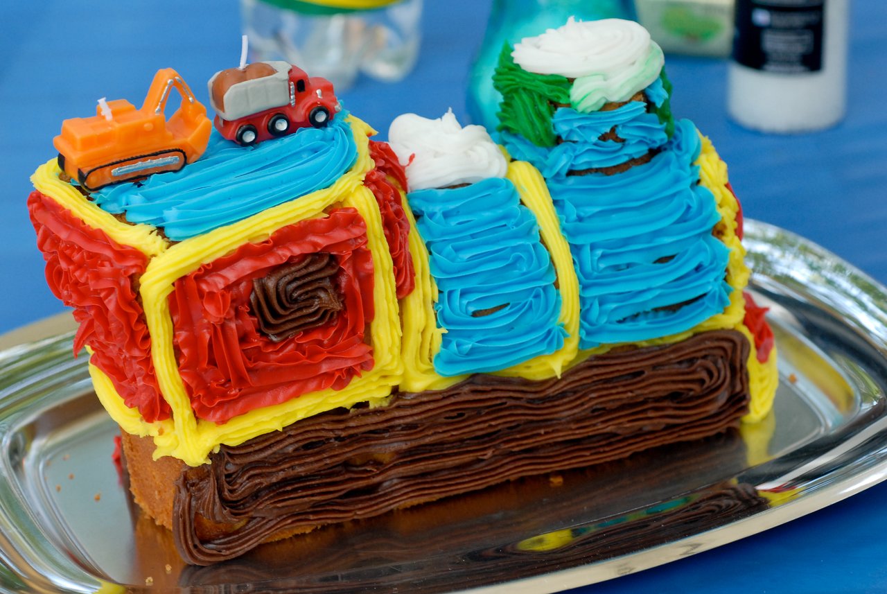 A colorful train-shaped birthday cake with toy construction vehicles on top, placed on a silver tray.