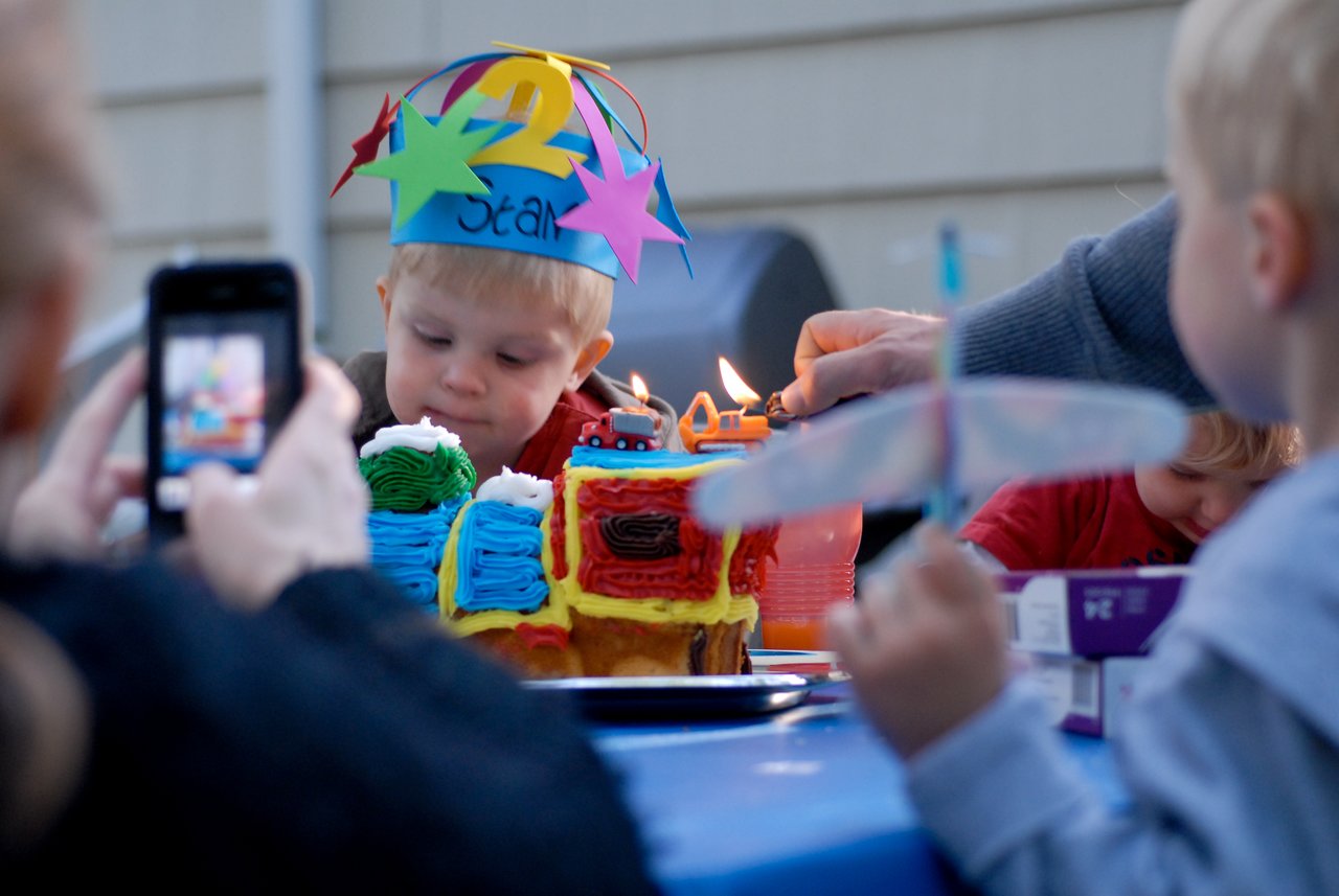 A young child wearing a colorful birthday hat watches as candles on a cake are being lit.