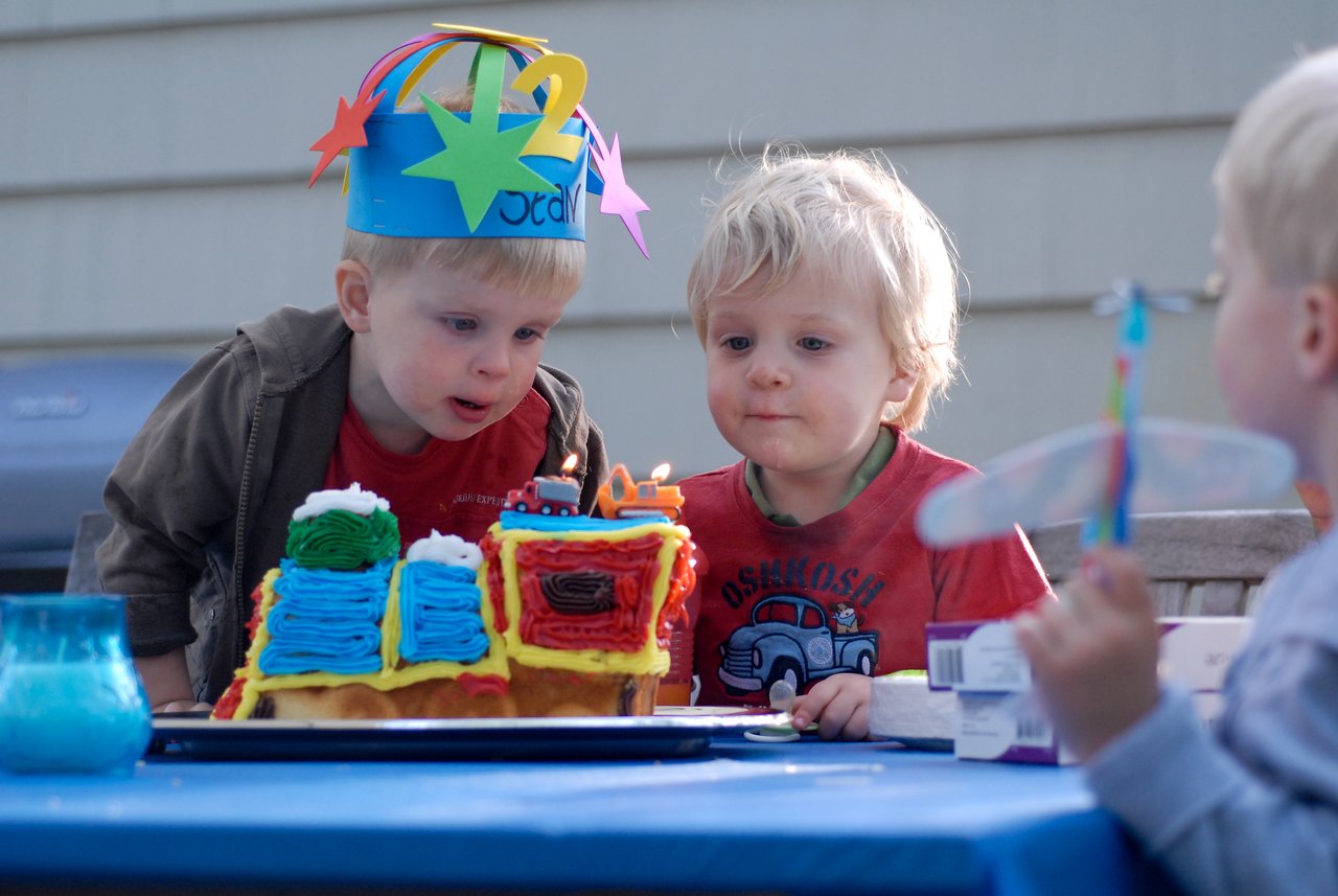 A child wearing a "2" birthday hat leans in to blow out candles on a colorful cake.