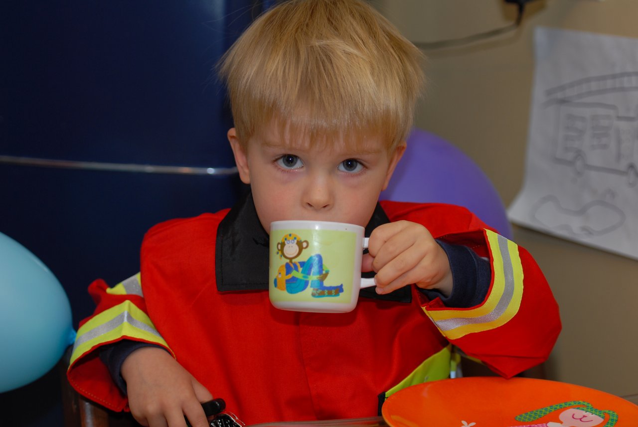 A young child in a firefighter costume drinks from a small cup while sitting at a table.