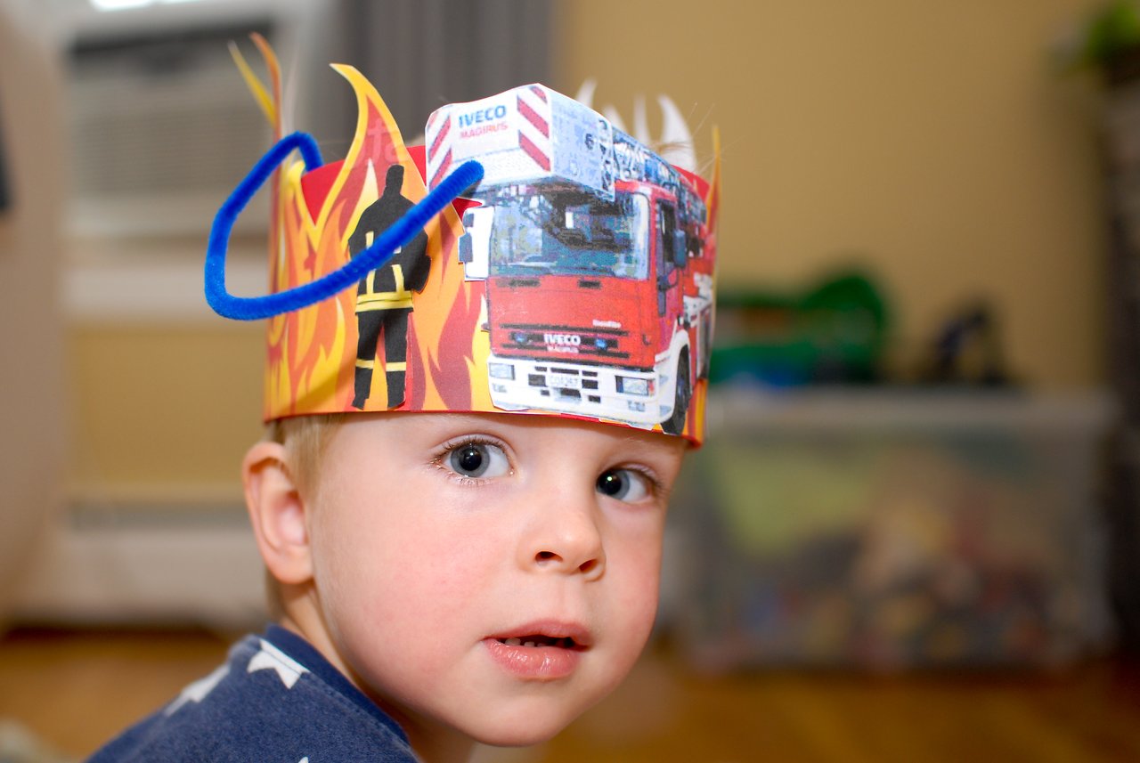 A young child wears a handmade firefighter-themed birthday crown with fire truck images and flame decorations.