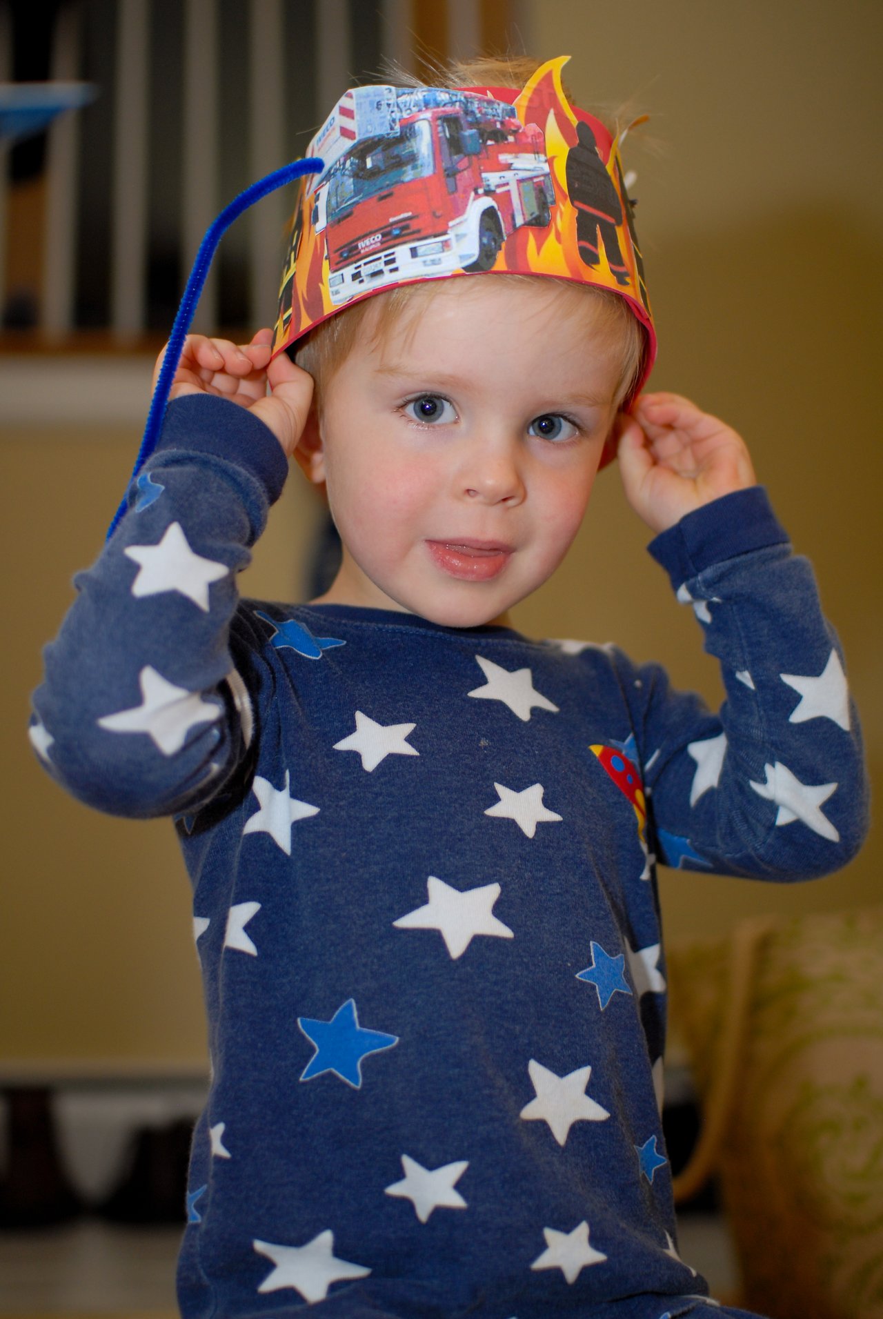 A young child wearing star-patterned pajamas adjusts a fire truck-themed birthday hat while looking at the camera.