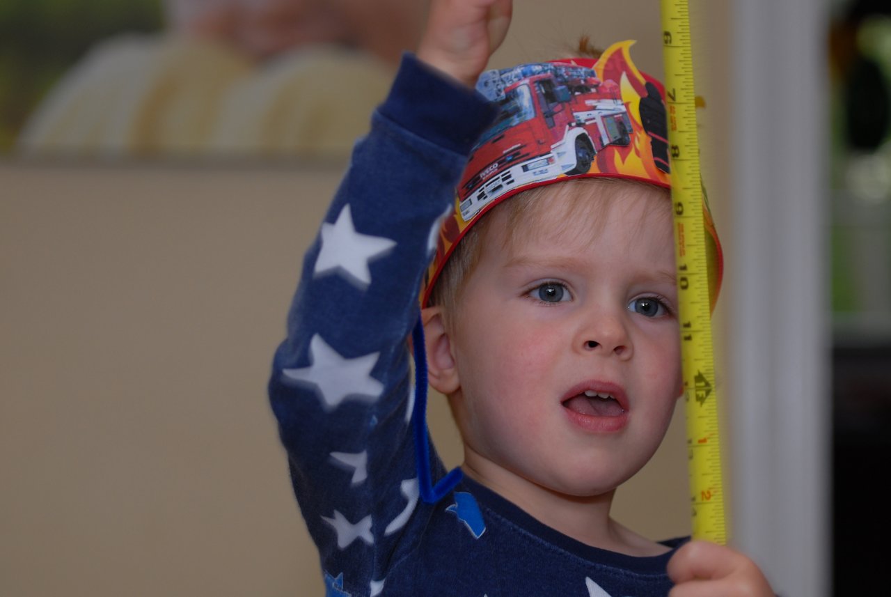 A young child wearing a fire truck-themed party hat holds up a measuring tape while looking up.