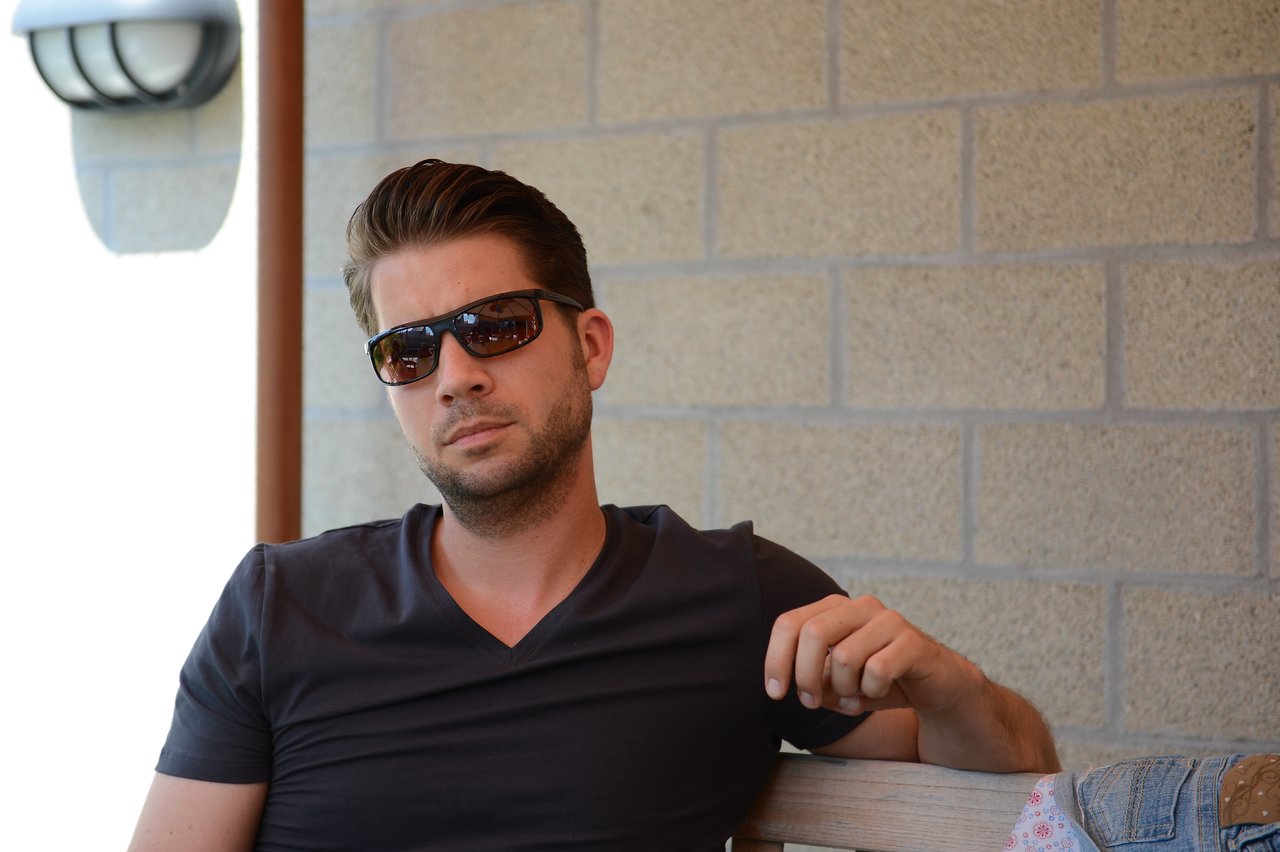 A man wearing sunglasses and a dark shirt sits on a bench, looking toward the camera with a neutral expression.