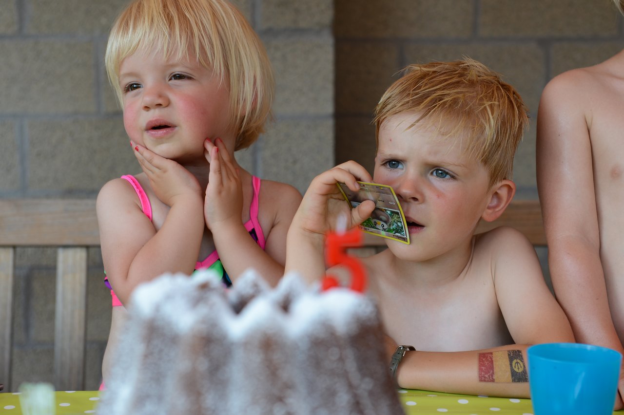 Two children in swimsuits sit at a table with a cake, one holding a card and looking focused.