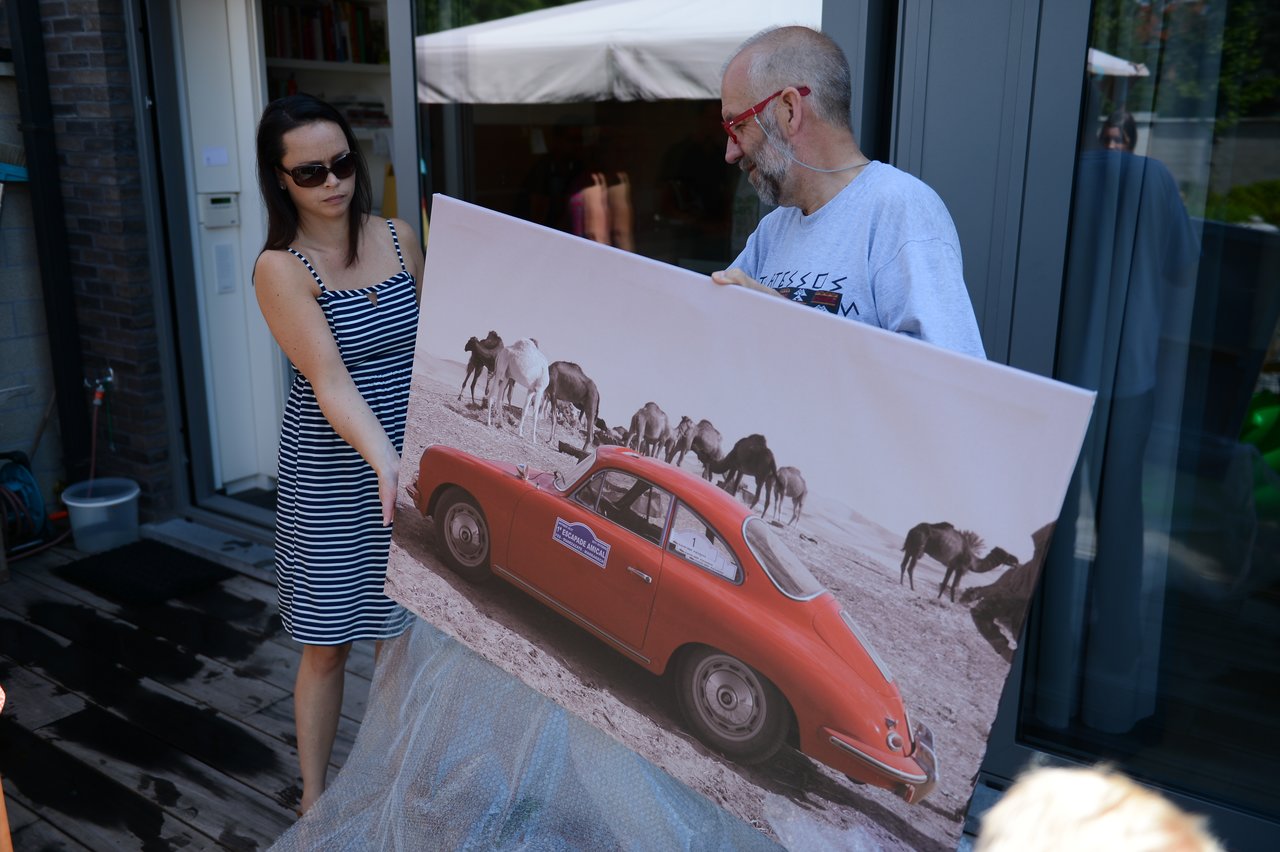 A woman and a man hold a large framed photo of a red car and camels in a desert.