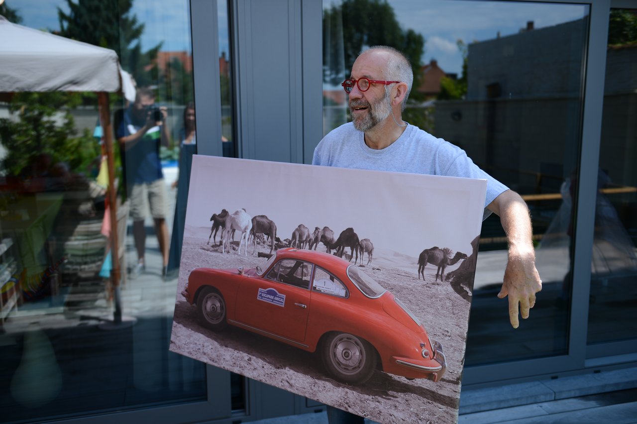 A man with red glasses holds a large framed photo of a red car and camels, smiling outside.