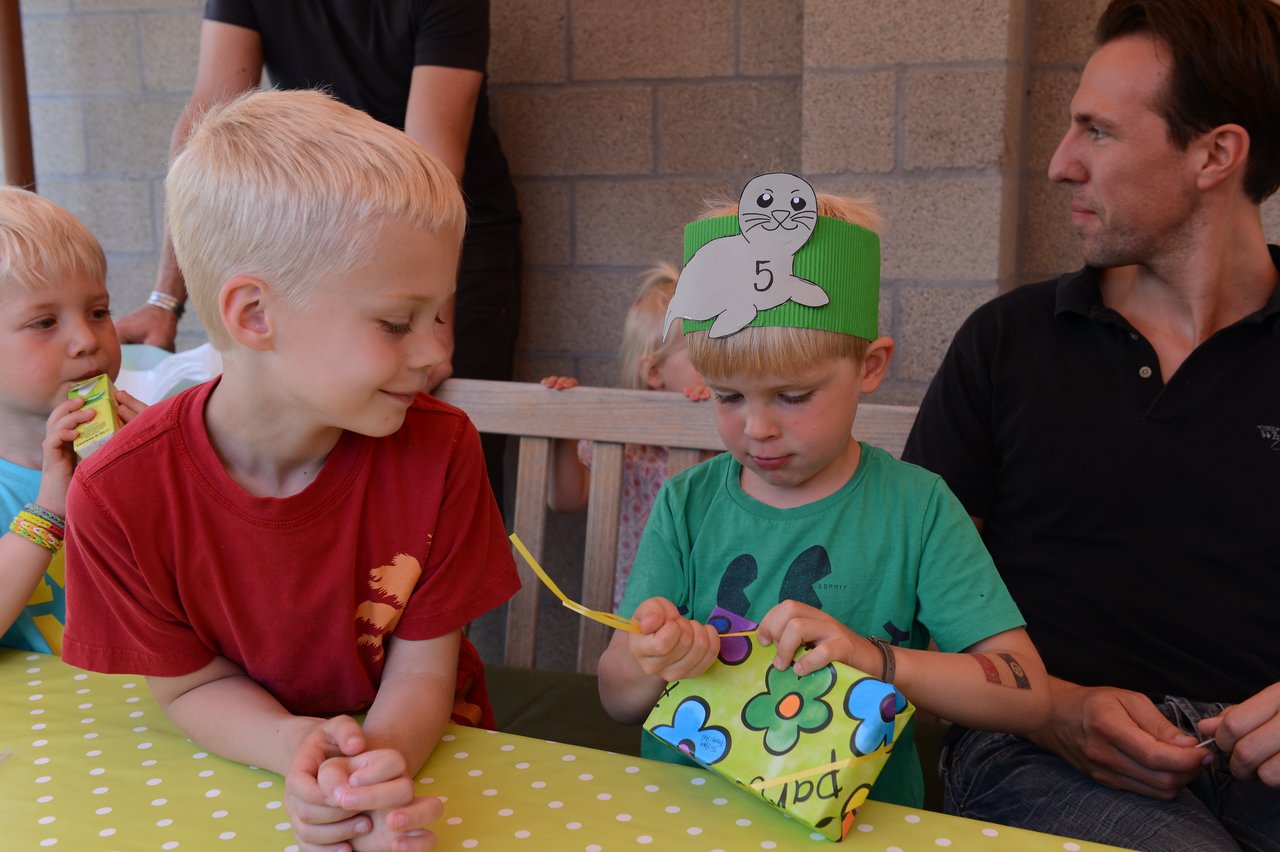 A young child wearing a seal-themed birthday hat opens a gift while others sit nearby at a party table.
