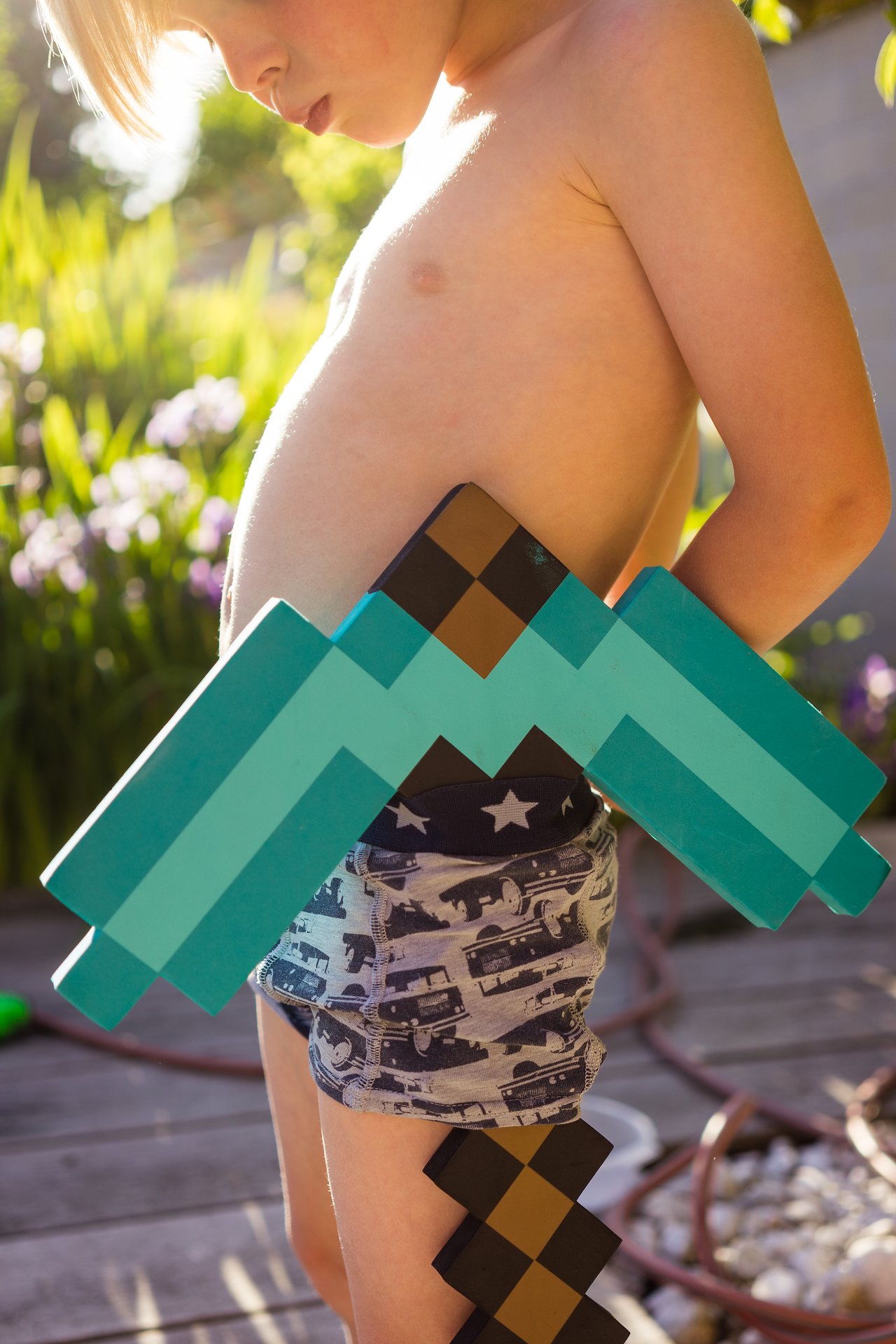 A shirtless child in patterned shorts holds a large pixelated toy axe outdoors in bright sunlight.