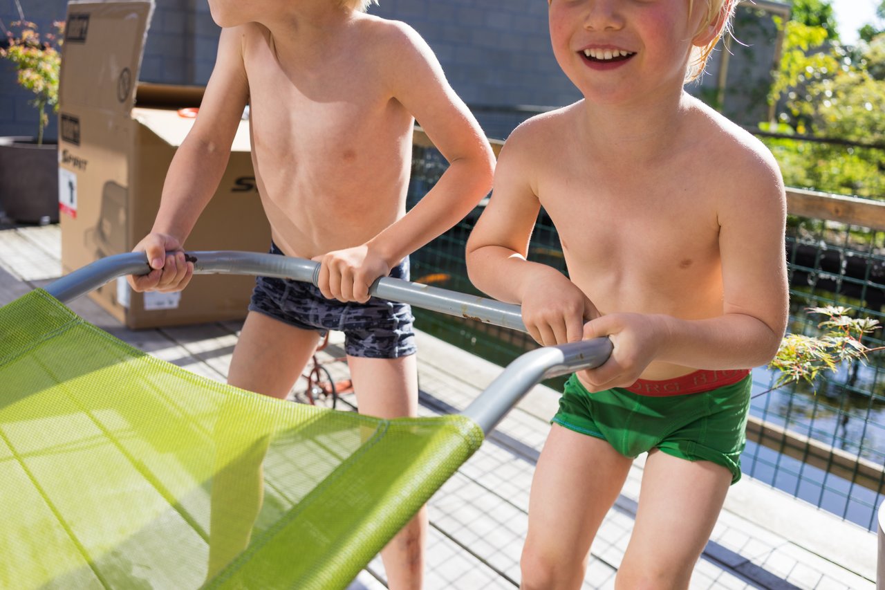 Two children in swimwear excitedly push a green lounge chair on a sunny outdoor deck.