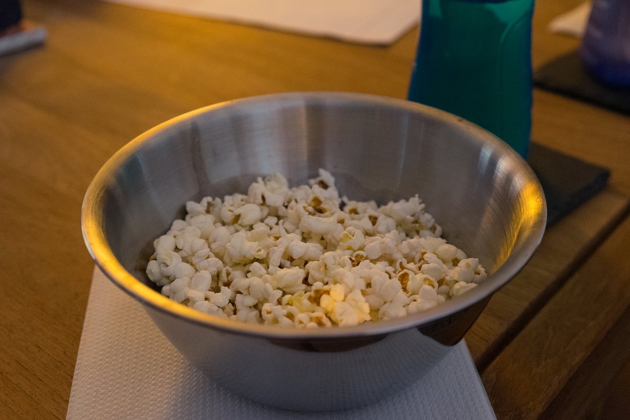 A metal bowl filled with popcorn sits on a wooden table, with drinks nearby.