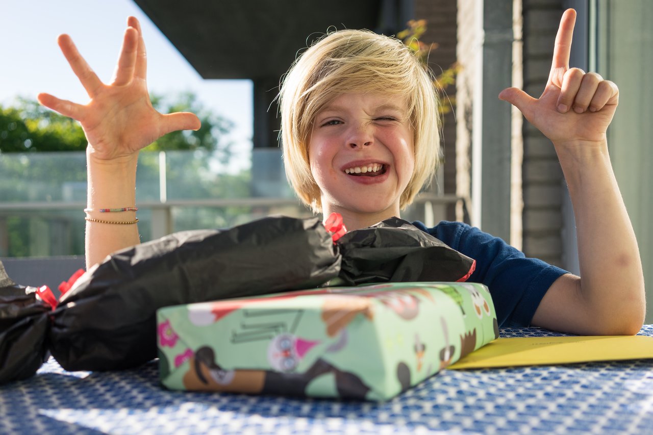 A child sits at a table with wrapped gifts, smiling and making playful hand gestures.