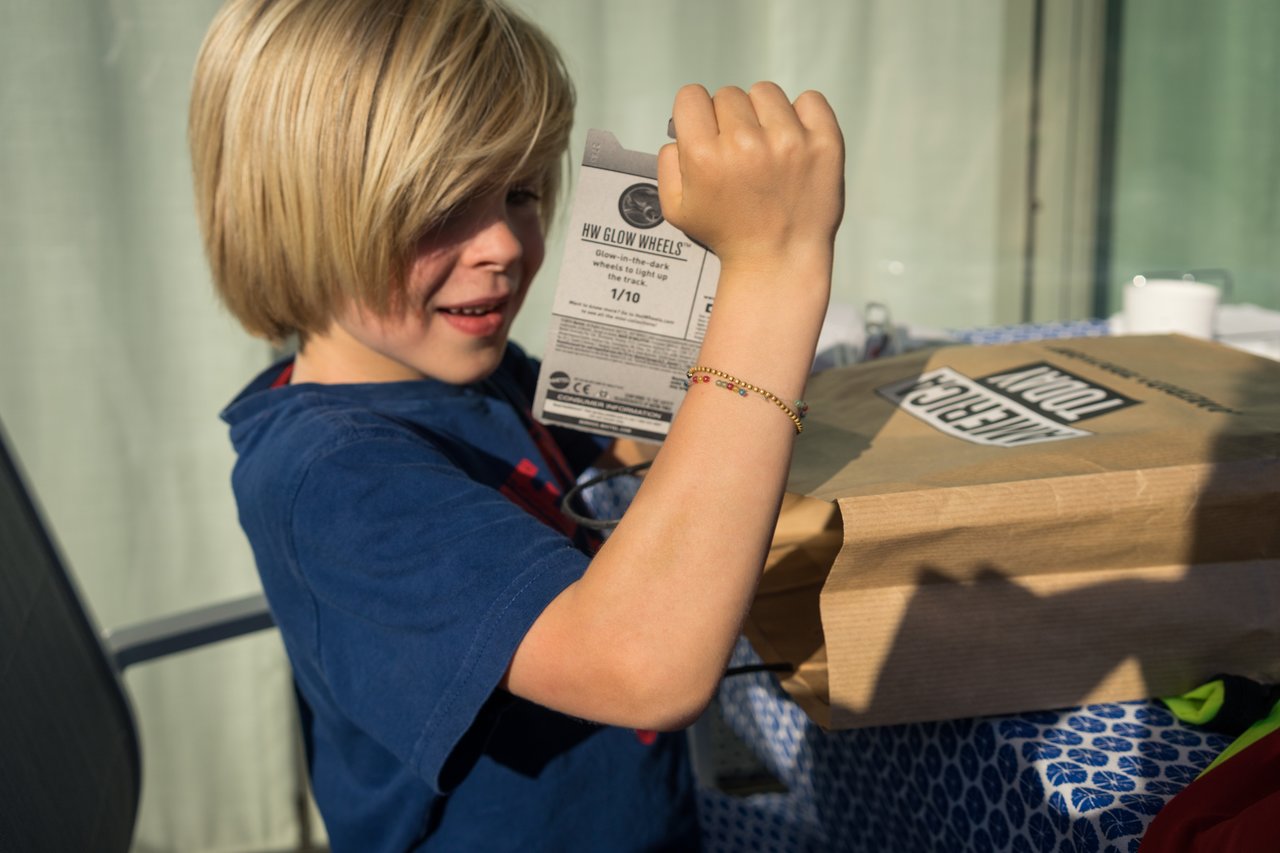 A child in a blue shirt excitedly examines a small toy package while sitting at a table with gifts.