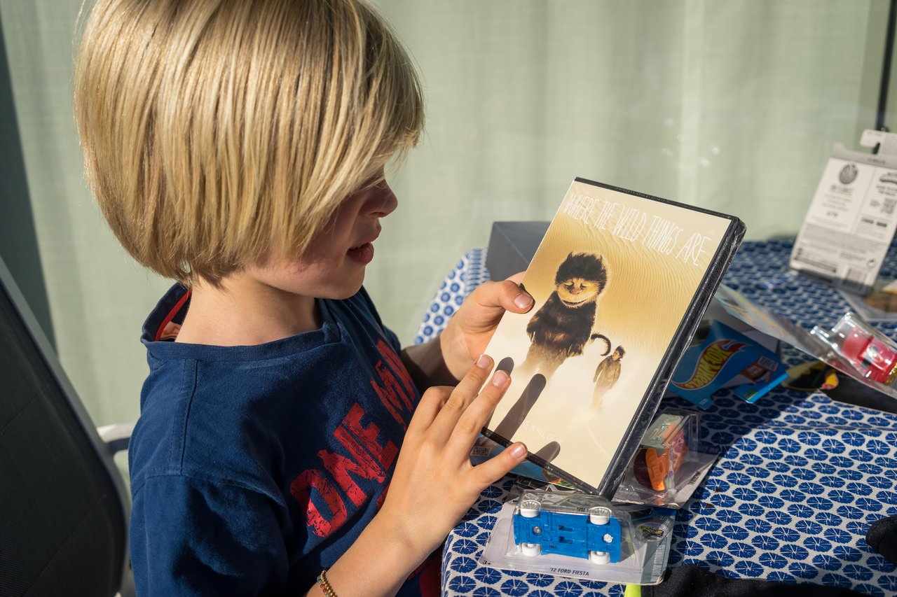 A child in a blue shirt holds a "Where the Wild Things Are" DVD while sitting at a table with toys.