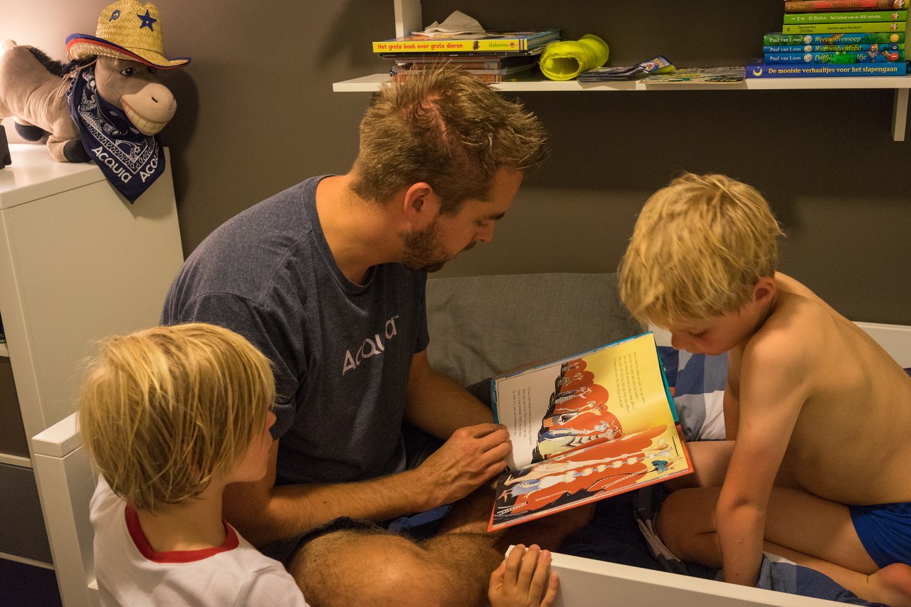A man sits on a bed reading a colorful storybook to two young children listening closely.