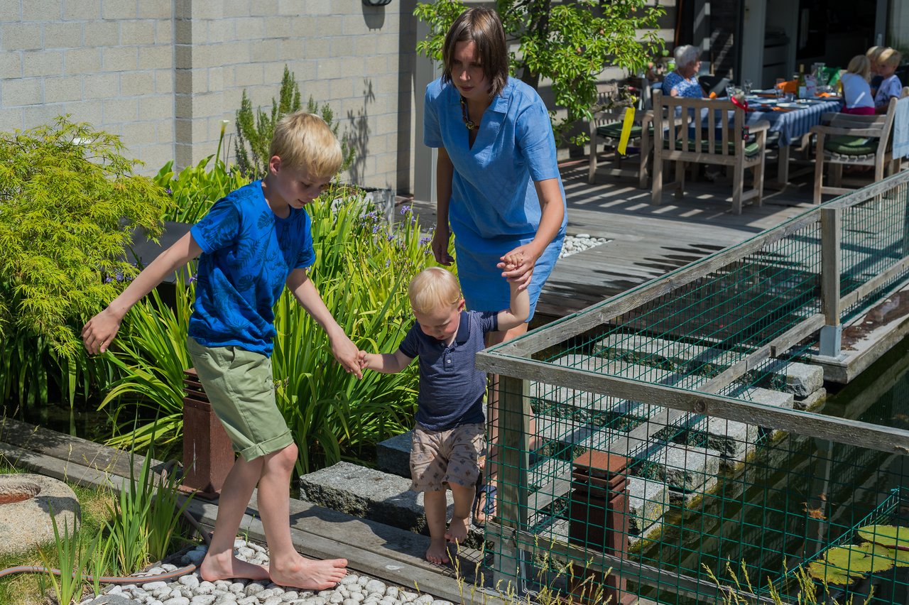 A woman helps two young children walk barefoot on a narrow path beside a small pond in a garden.