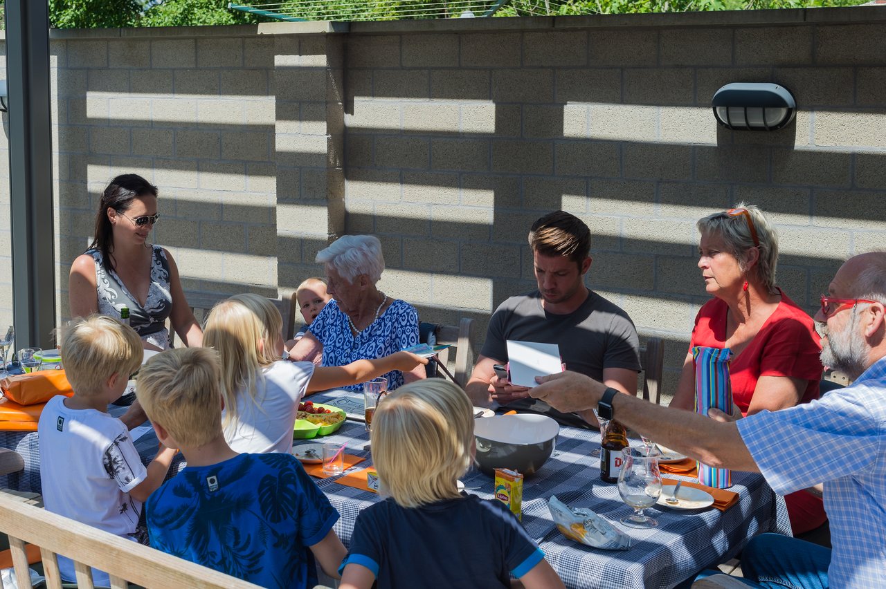 A group of people, including children and adults, gather around a table outdoors, sharing food and conversation.