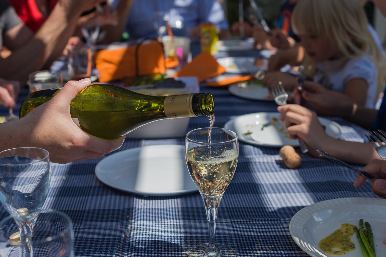 A person pours white wine into a glass at an outdoor gathering with people eating at a table.
