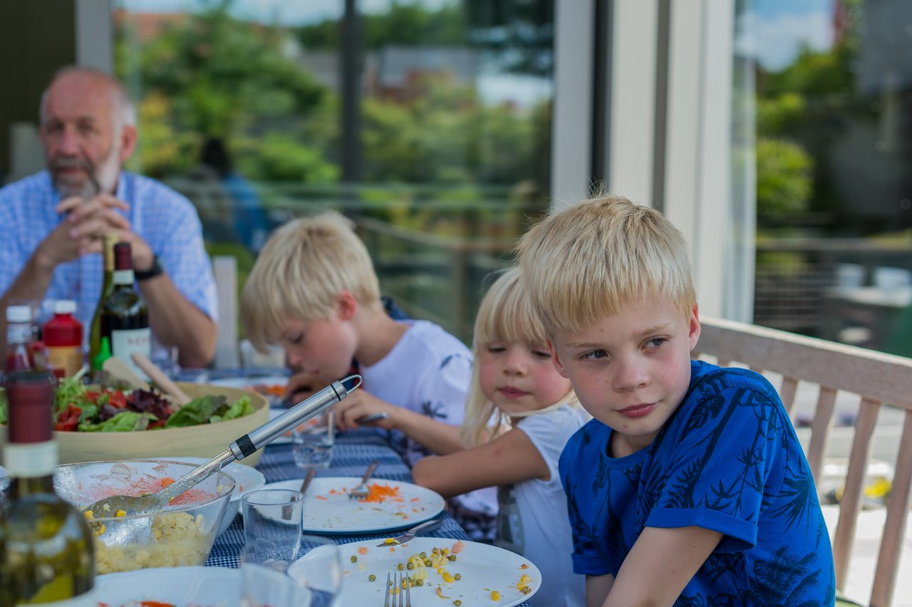 Children and an older man sit at an outdoor table with food, drinks, and salad during a barbecue gathering.