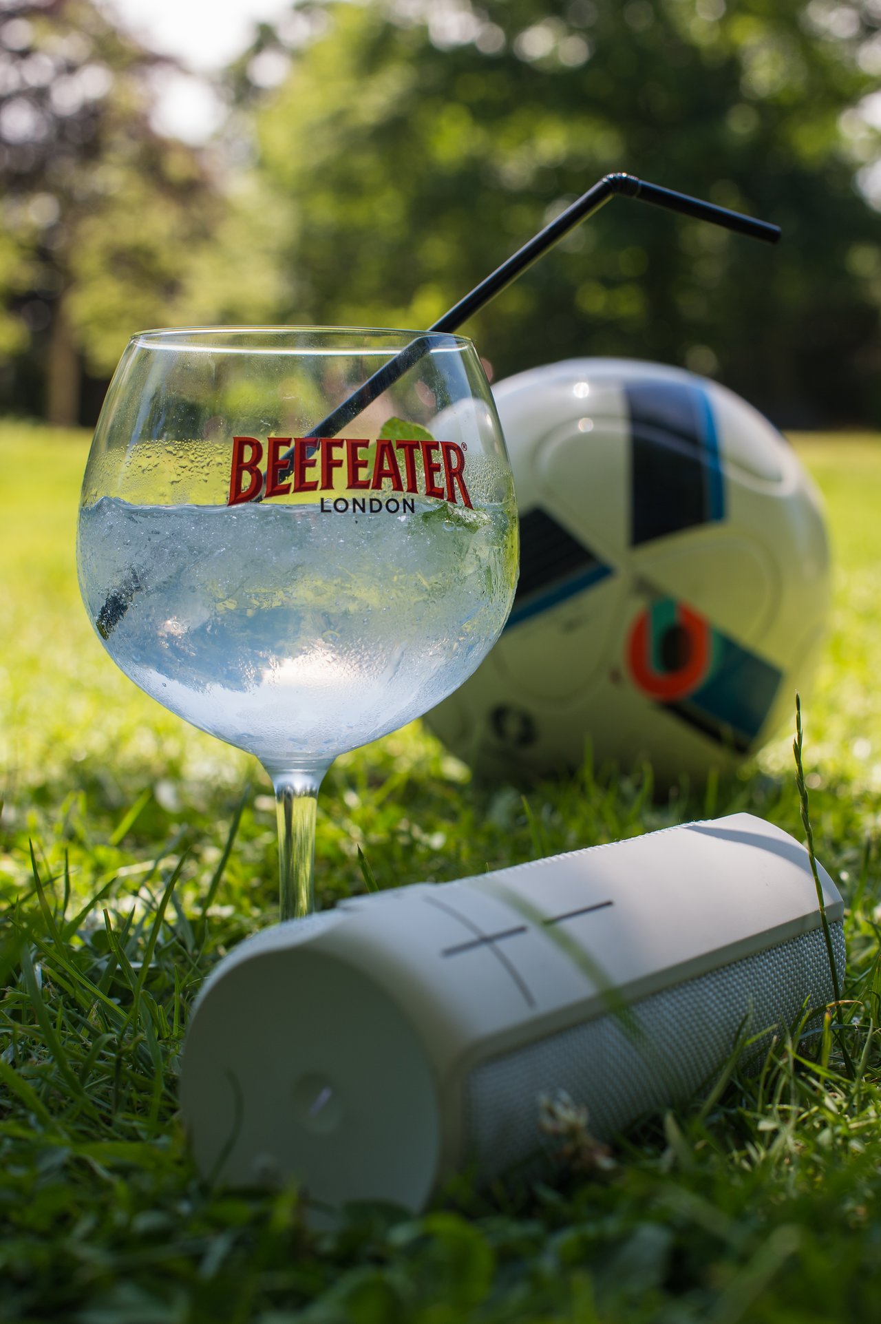 A glass with a drink, a speaker, and a soccer ball rest on the grass in a park.