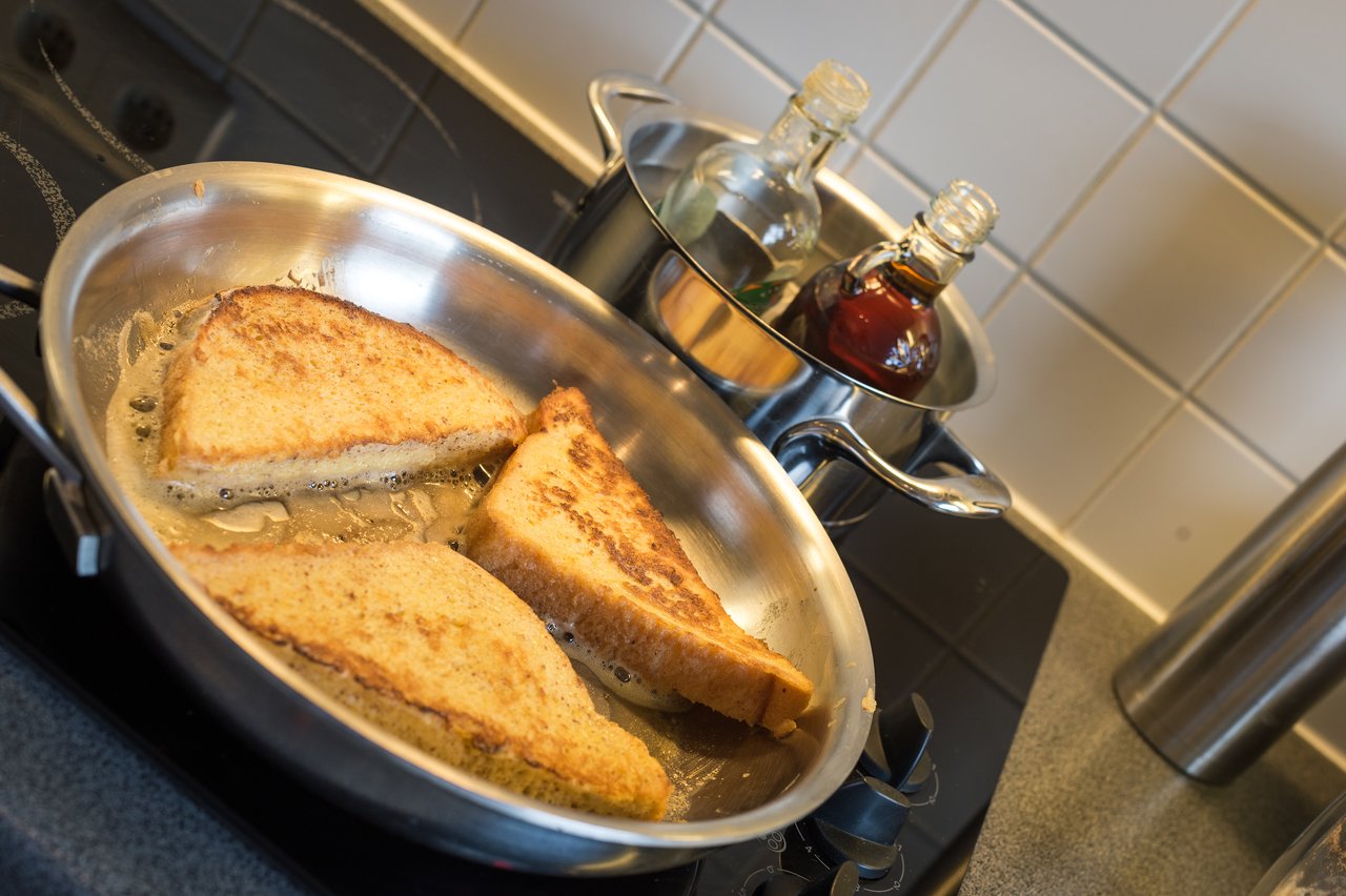 Three slices of French toast cooking in a frying pan on the stove, with syrup bottles nearby.