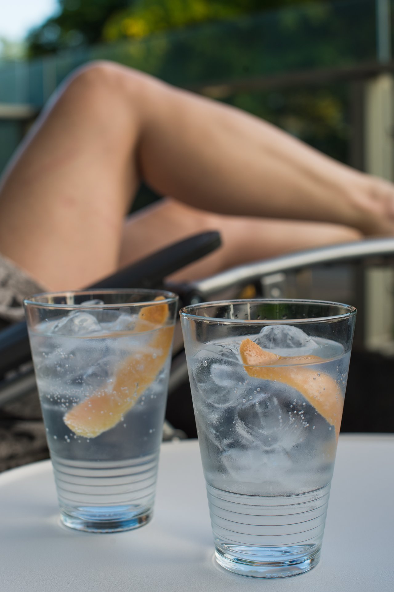 Two glasses of gin and tonic with ice and citrus peel on a table, with legs resting nearby.