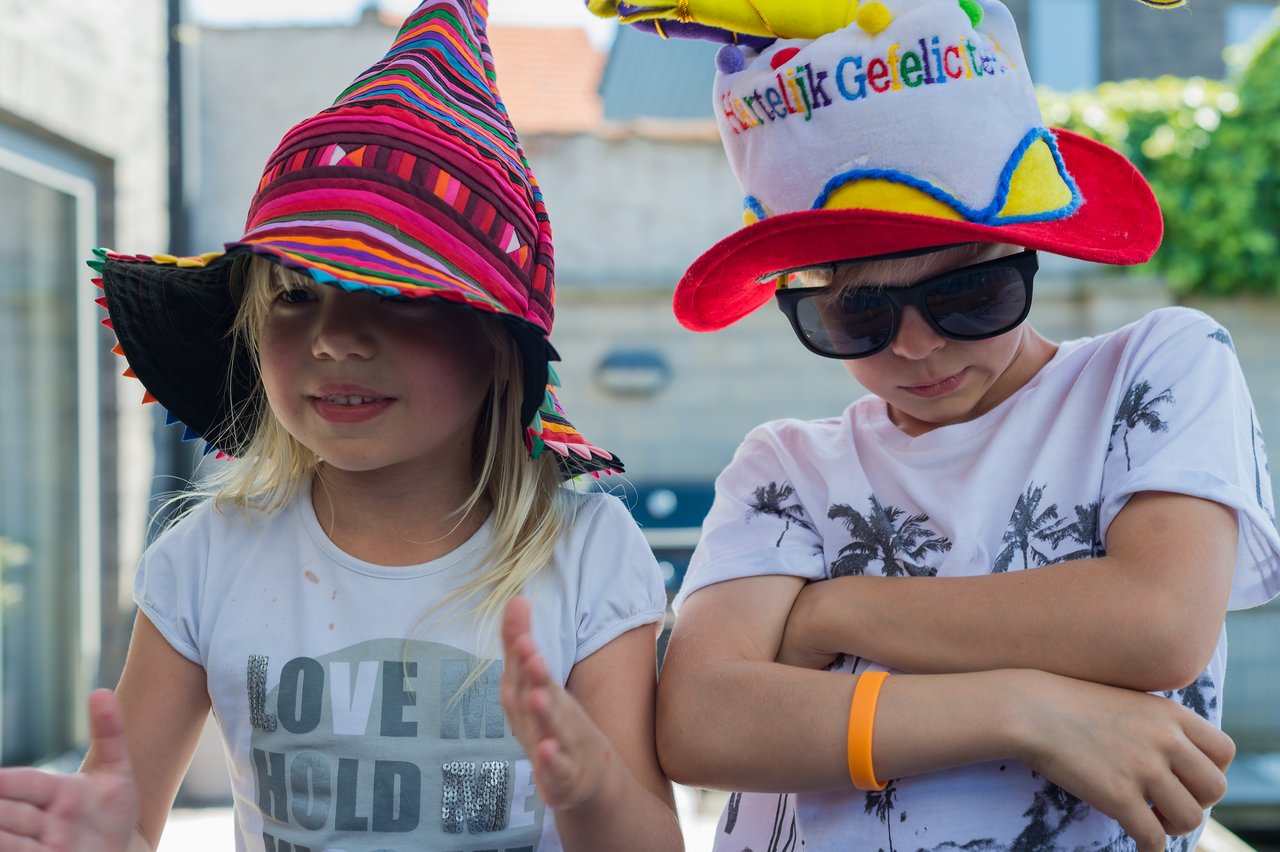 Two children wearing colorful hats pose playfully, one clapping and the other crossing their arms with sunglasses on.