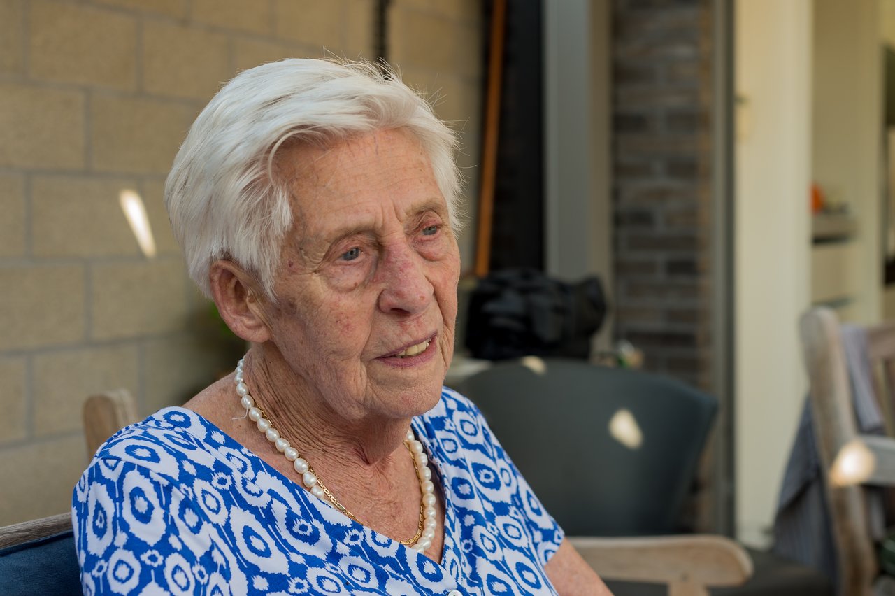 An elderly woman with short white hair sits outside, wearing a blue patterned dress and a pearl necklace.