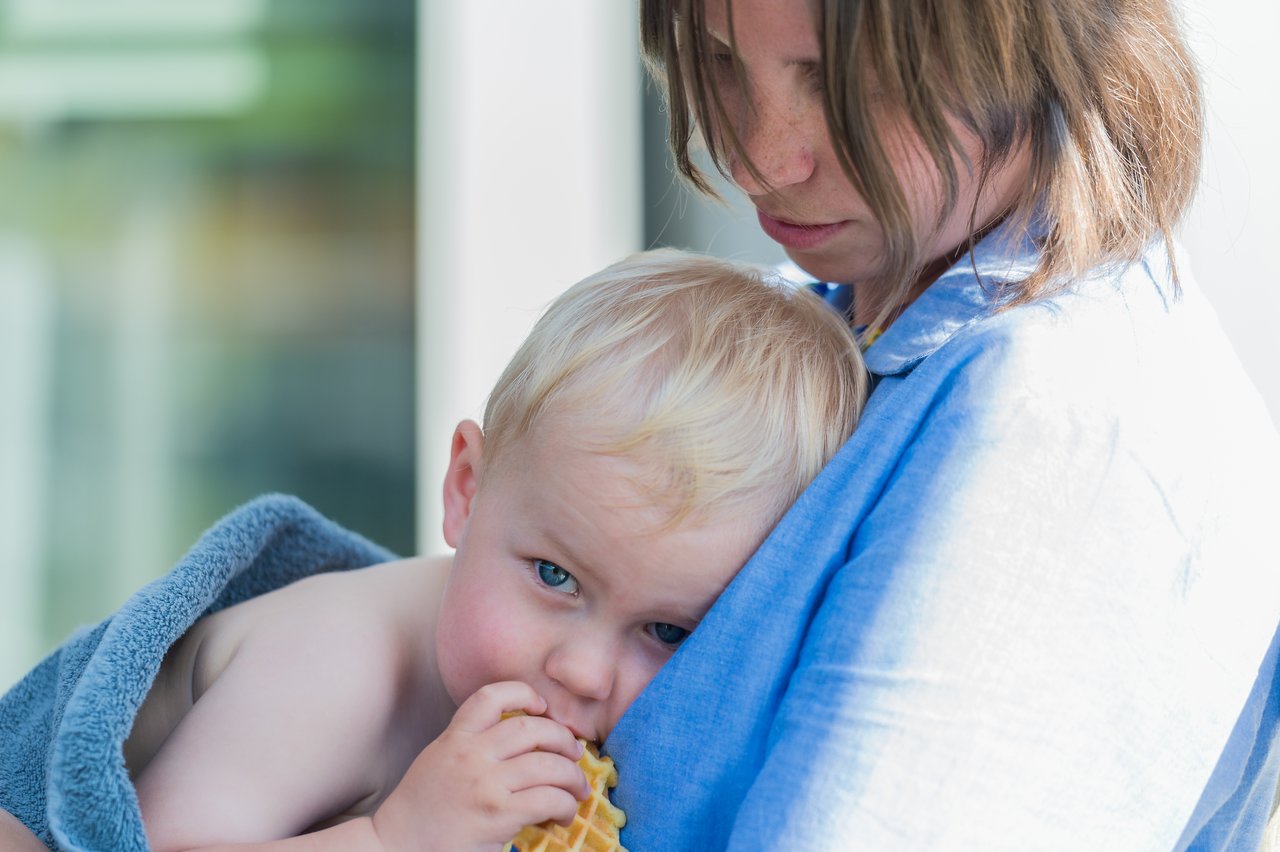 A young child wrapped in a towel eats a waffle while being held by an adult wearing a blue shirt.