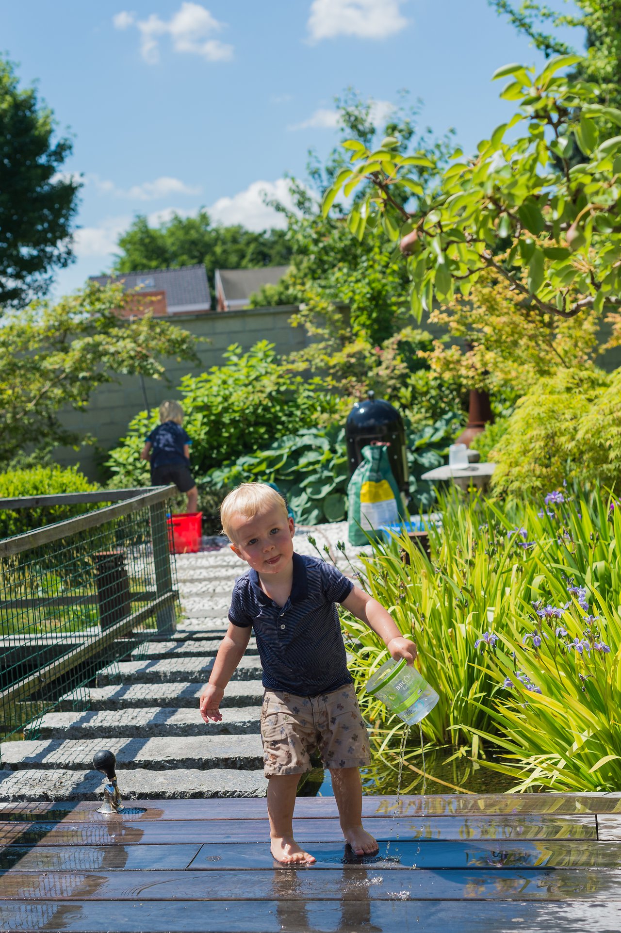 A young child with wet clothes holds a plastic container, playing near water on a sunny day.