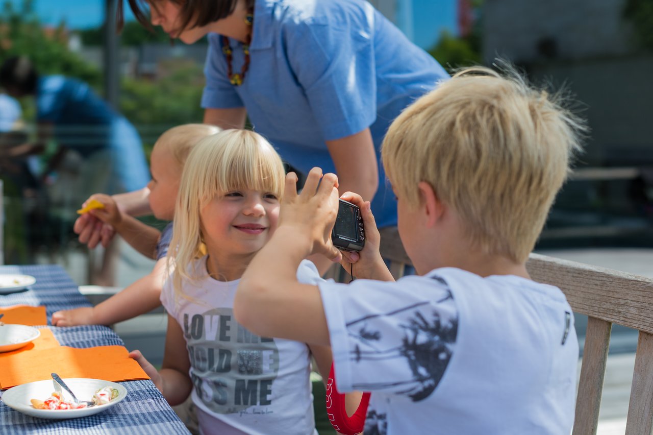 A child holds a small camera, taking a photo of a smiling girl sitting at a table during a gathering.