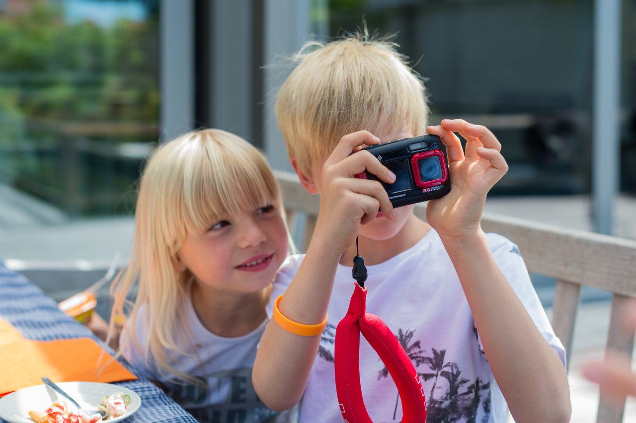 A child holds a camera and takes a photo while another child watches and smiles.