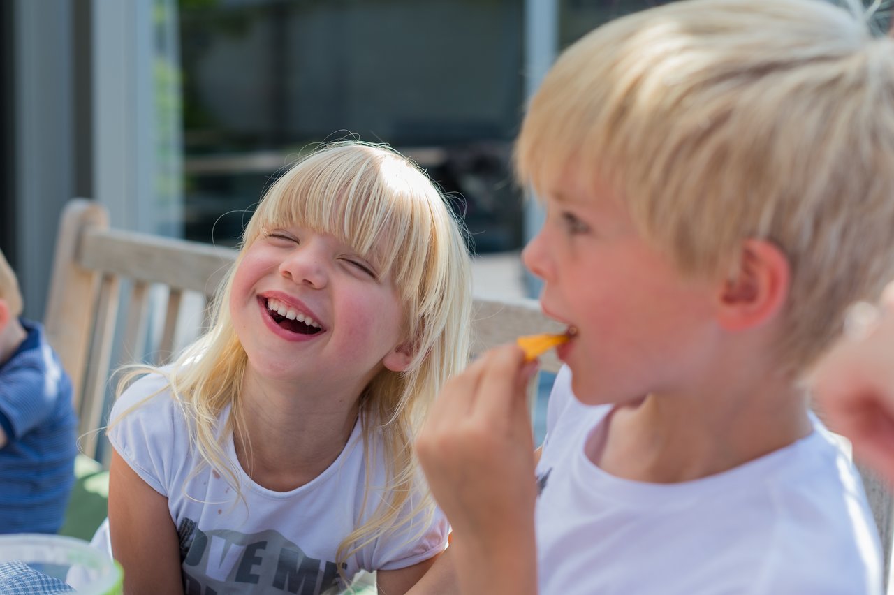 A young girl laughs while sitting next to a boy eating a snack outdoors.