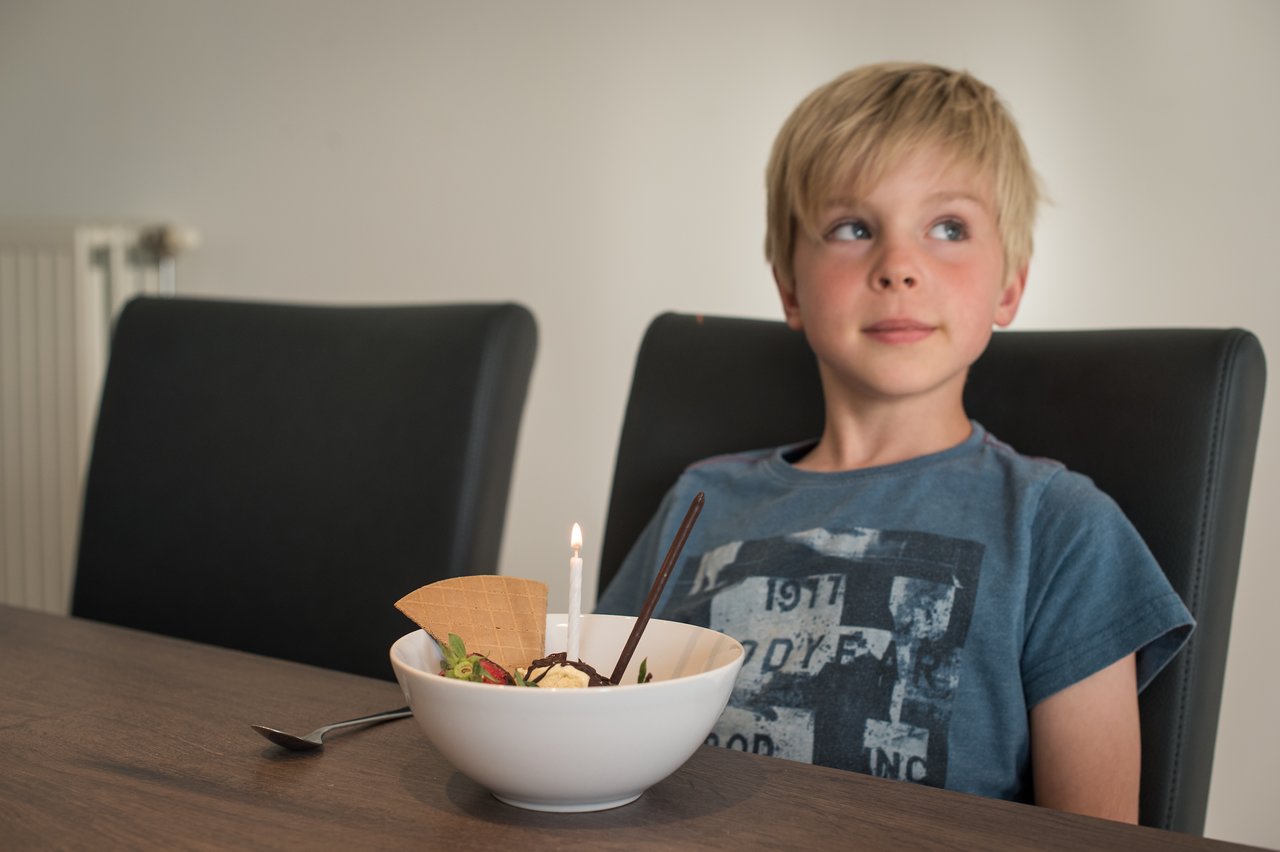 A boy sits at a table with a dessert bowl holding a lit birthday candle, looking to the side.