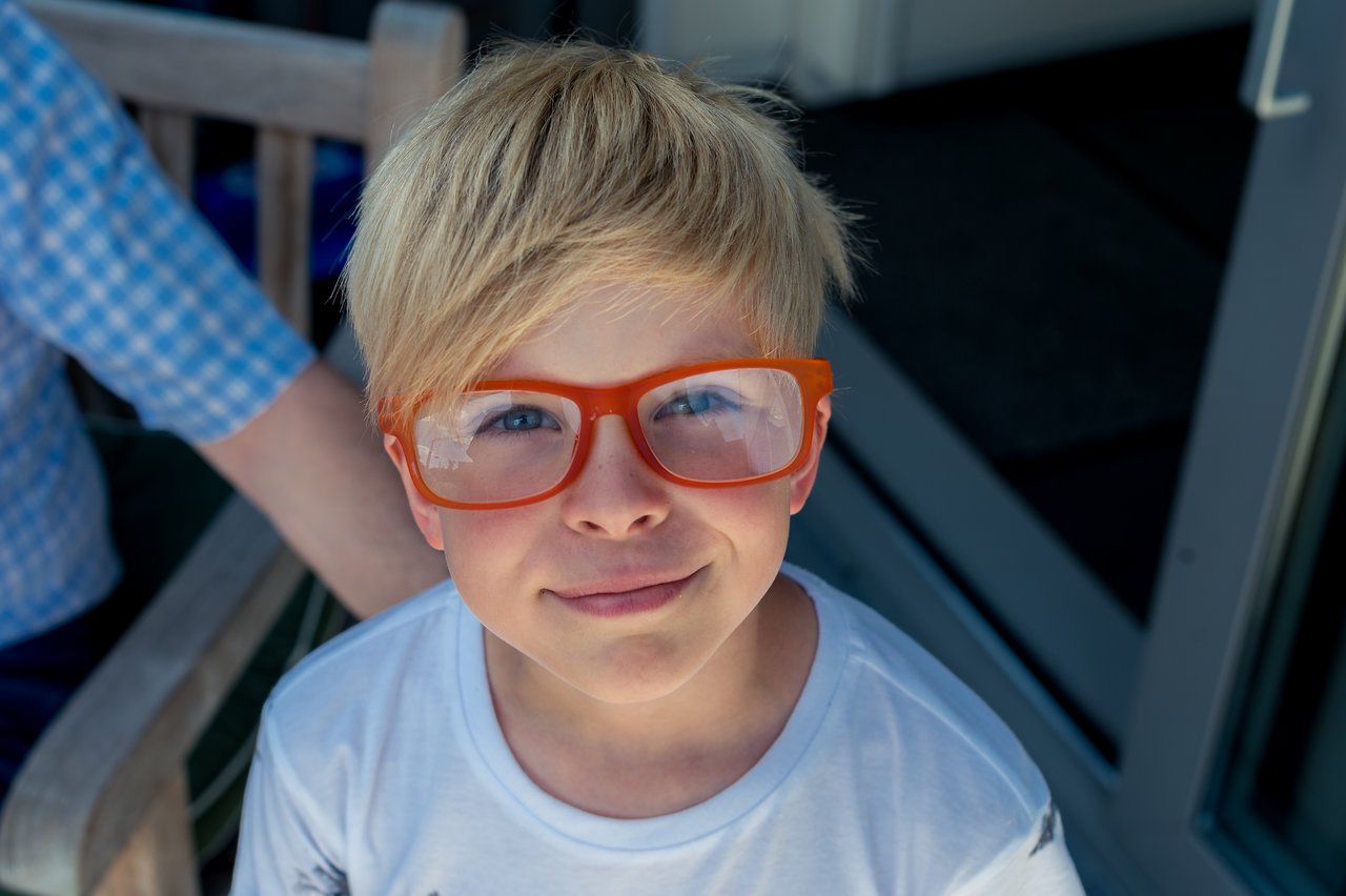 A young boy with blond hair wears oversized orange glasses and smiles at the camera.