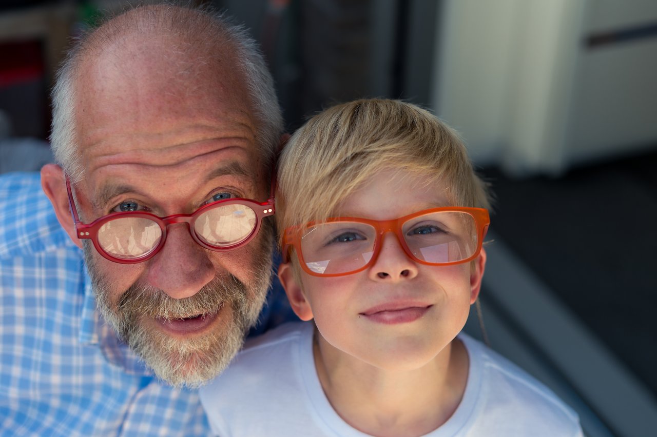 A young boy and an older man smile while wearing similar glasses, looking directly at the camera.