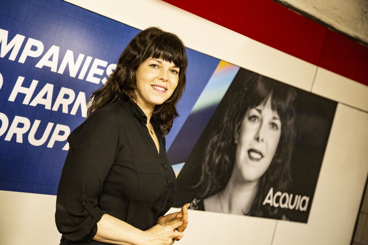 A woman in a black shirt stands in front of a wall with her photo and the word "Acquia.
