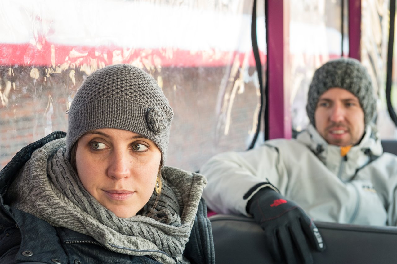 Two people in warm clothing sit inside a Boston Duck Tour vehicle, one looking away while the other watches.