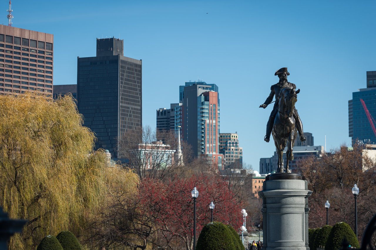 Equestrian statue on a pedestal in a park, with modern buildings in the background under a clear blue sky.