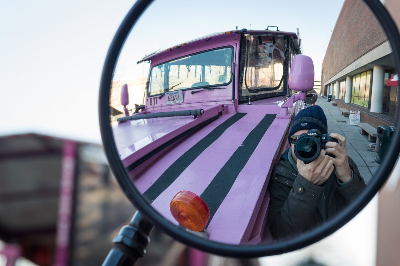 A photographer captures their reflection in the side mirror of a pink Boston Duck Tour vehicle.